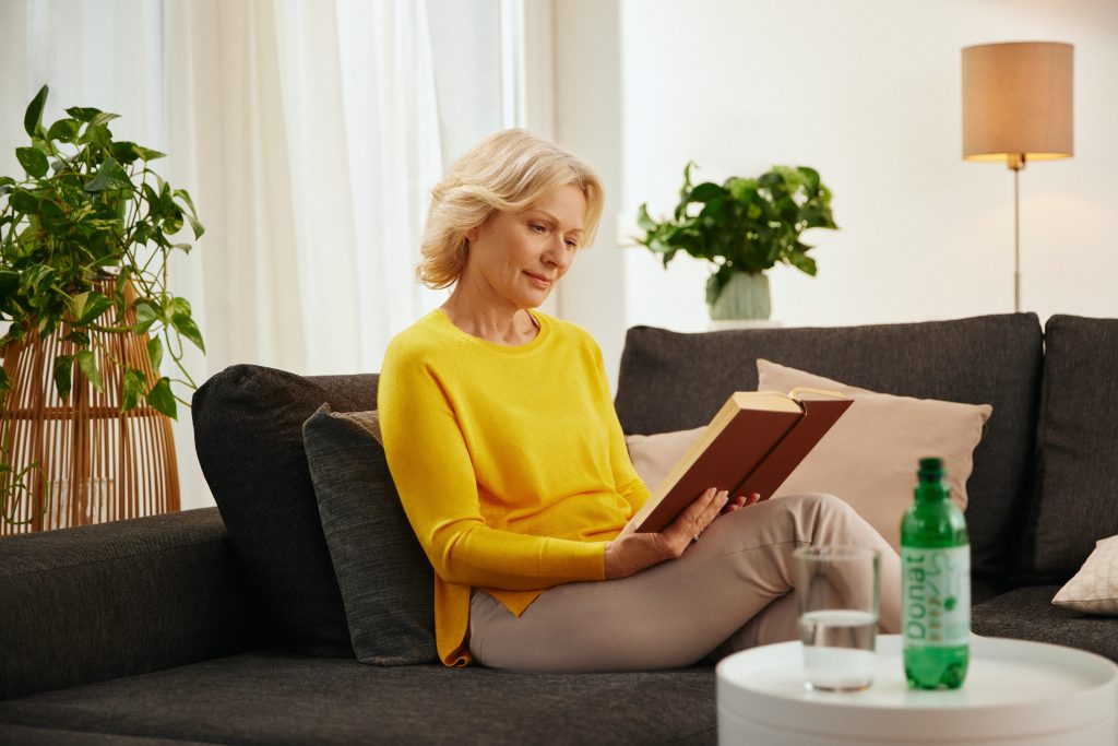 An elderly woman is sitting on the couch reading a book and drinking Donat mineral water.