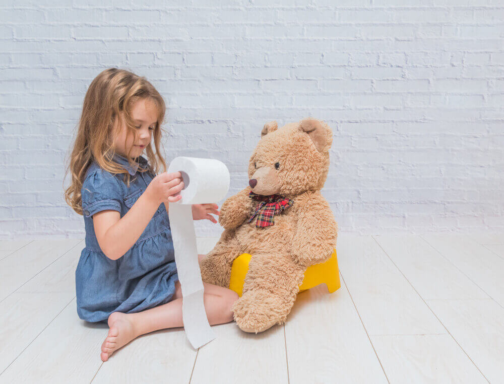 Ein Mädchen sitzt auf dem Boden und spielt mit einer Rolle Toilettenpapier, neben ihr sitzt ein Teddybär auf dem Nachttopf.